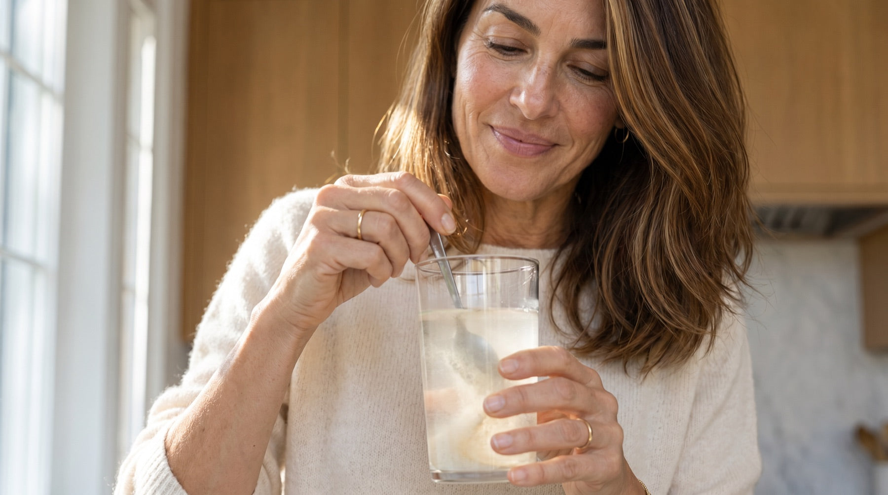 The same woman stirring her cloudy electrolyte-and-creatine drink in a sunlit afternoon kitchen.